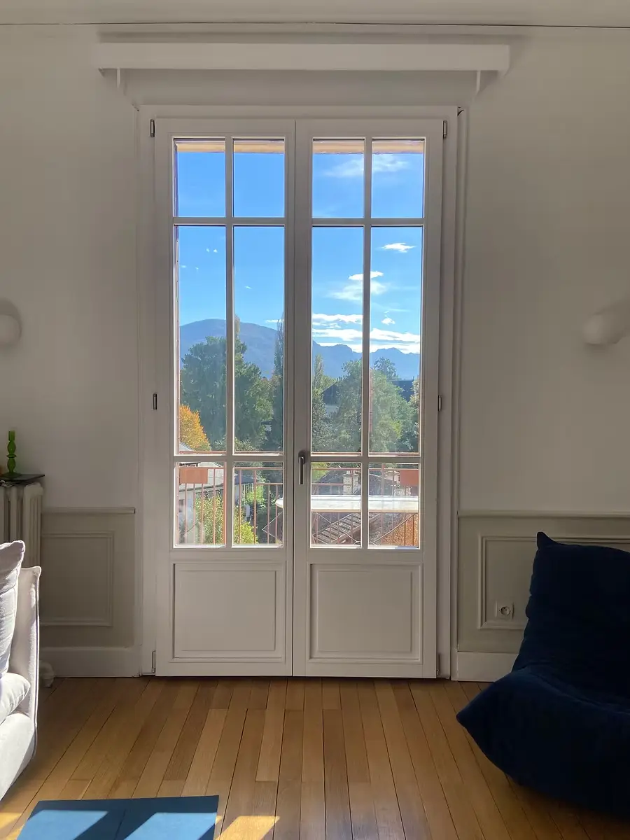 Porte-fenêtre blanche à petits bois, sous une corniche moulurée, ouverte sur la montagne.