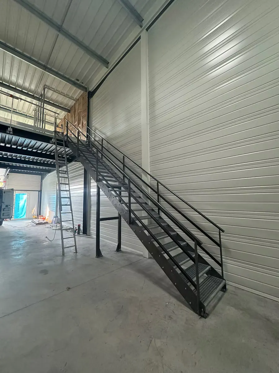 Escalier métallique droit avec garde-corps, montant vers une mezzanine dans un bâtiment en bardage.
