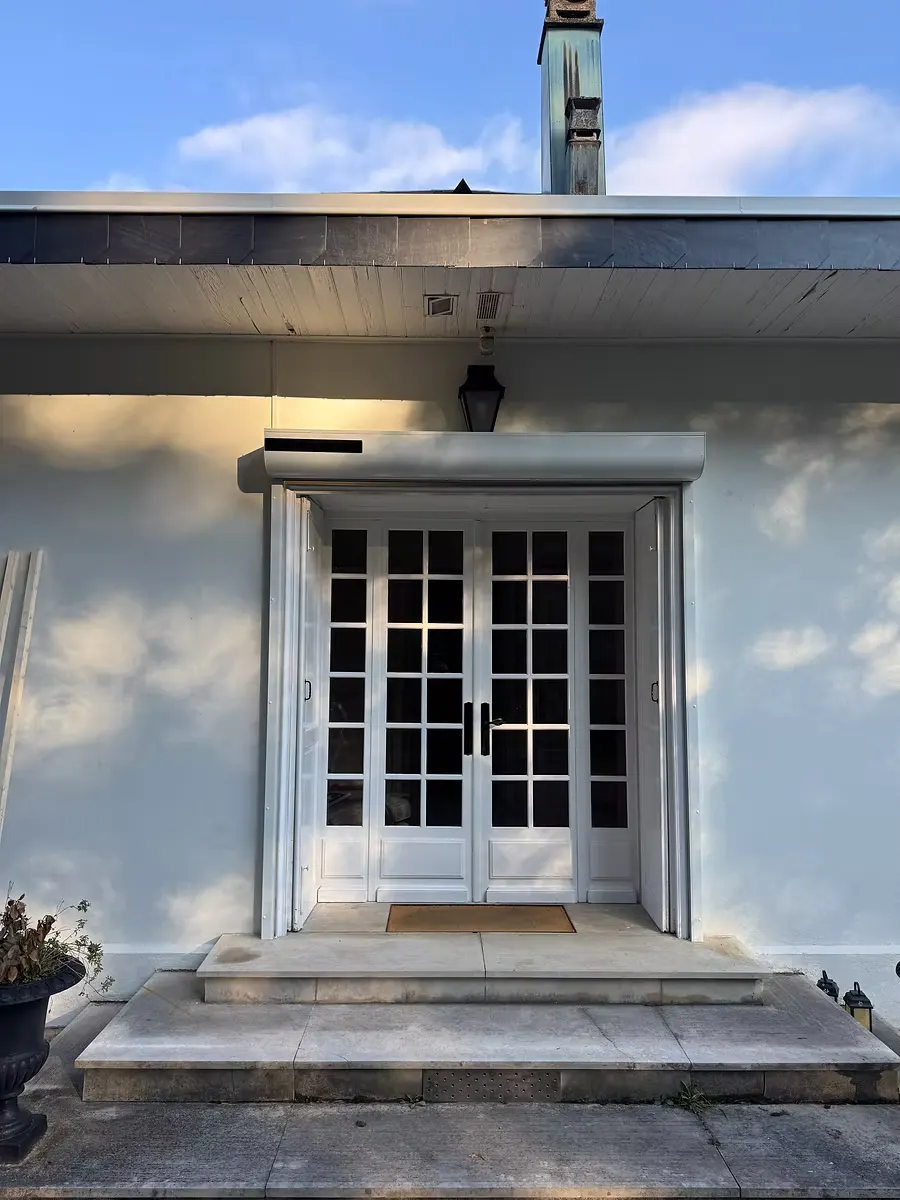 Porte-fenêtre double à petits bois au centre d’une façade blanche, sous un auvent.