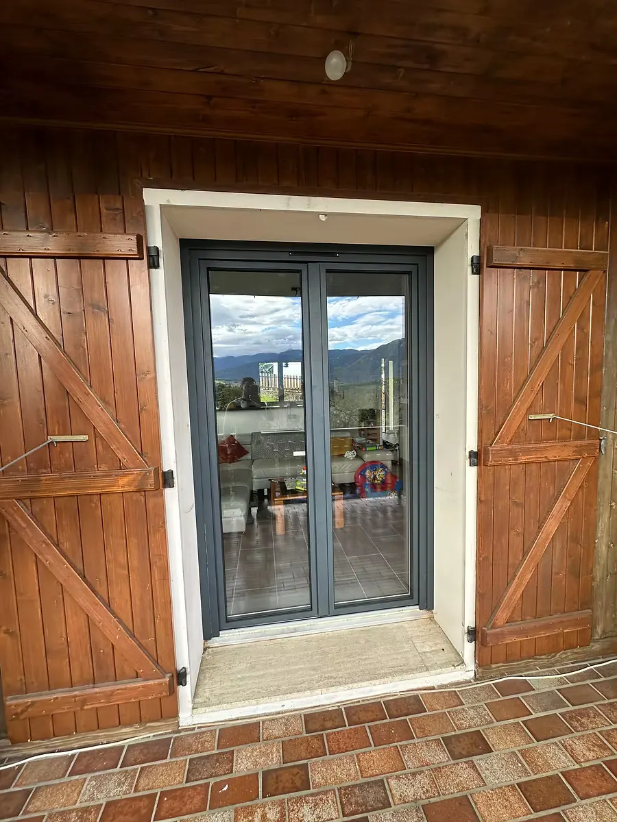 Porte-fenêtre gris anthracite encadrée de volets en bois, sol en tomettes.