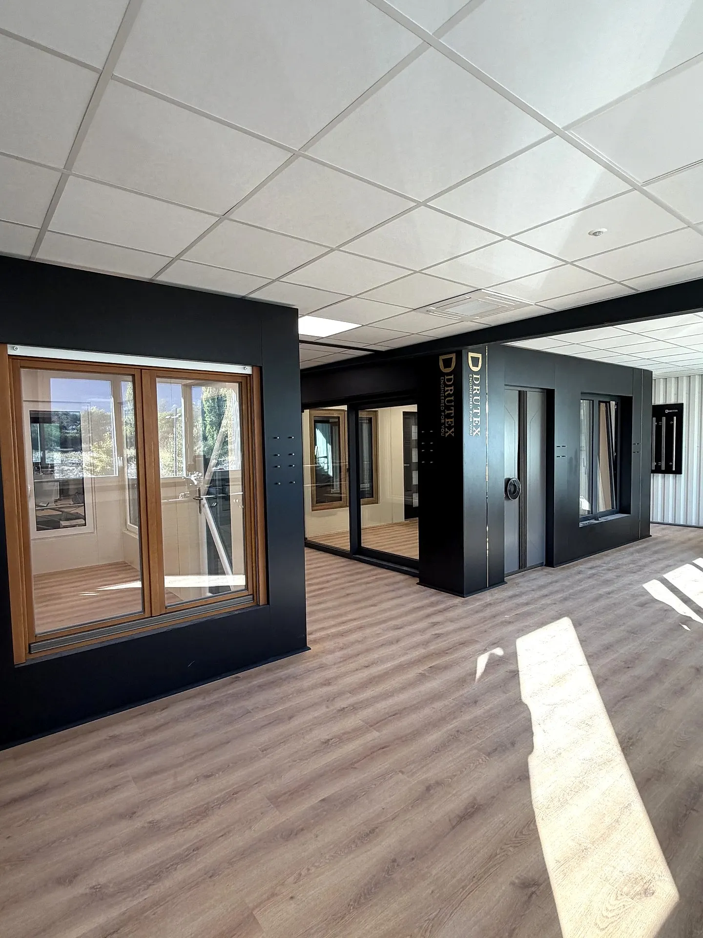 Le showroom SMV Habitat à Méry : cloisons noires où sont posées une porte-fenêtre bois, des fenêtres et des portes DRUTEX, sur un parquet clair.