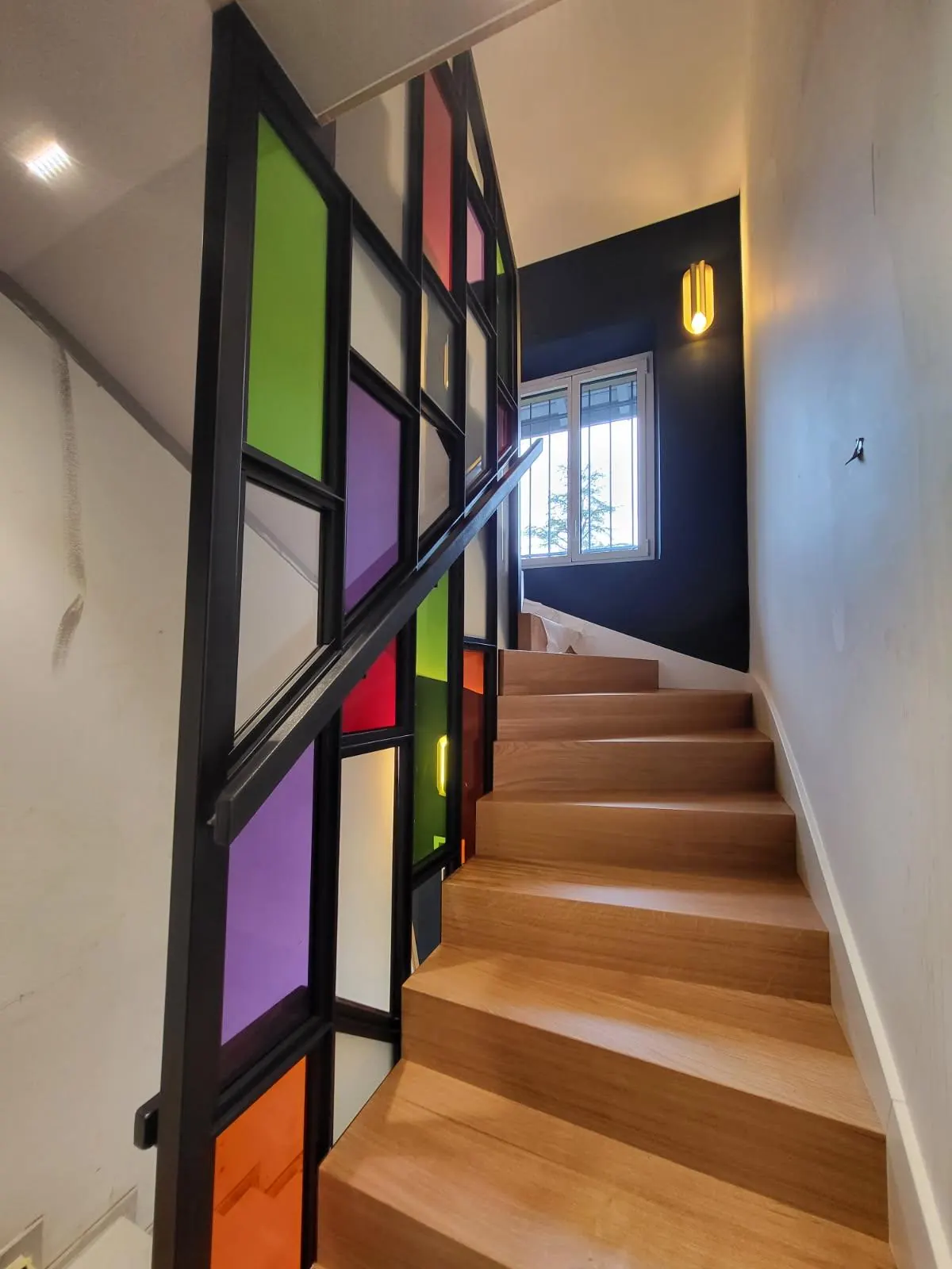 Escalier en chêne bordé d’une verrière en acier noir aux vitrages de couleur, vert, violet, rouge et orange, avec sa main courante.